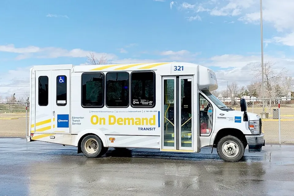 White minibus with yellow "On-Demand" logo parked in an asphalt lot with grass field and dry fall trees in background in Edmonton, Canada.